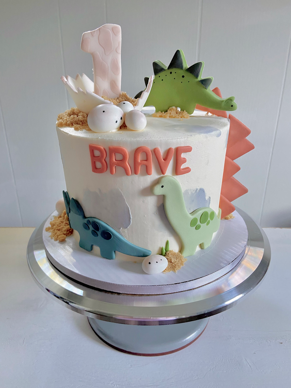 Cake with fondant dinosaurs, a one topper, speckled fondant dino eggs and brown sugar sand.