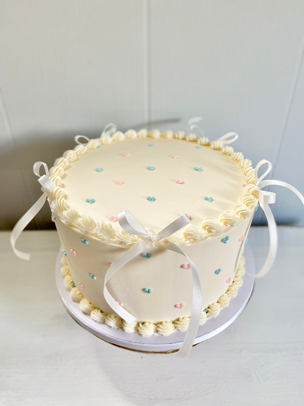 Round cake with white frosting tiny blue and pink heart polka dots. Ribbon bows in the top border.