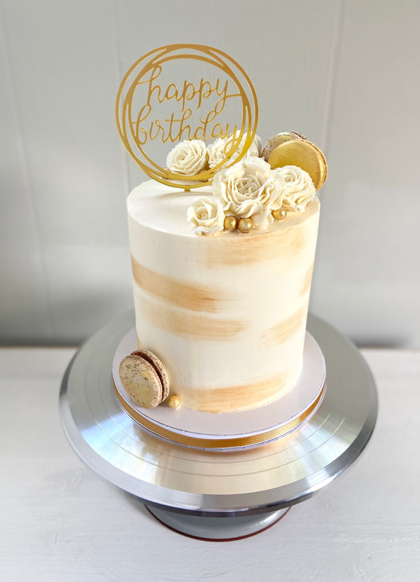 White and gold cake with buttercream flowers, macarons, and a gold happy birthday topper.