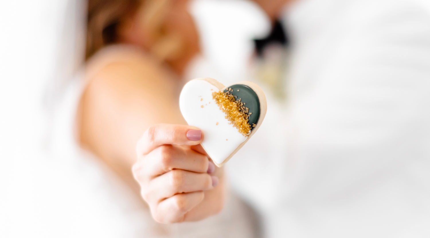 Closeup of bride's hand holding heart shaped cookie decorated with white and green icing.