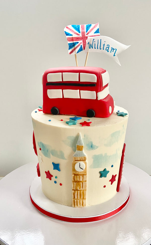 Single tier London themed cake with fondant double-decker bus topper and British flag. Sides have fondant Big Ben, telephone booth, and red white and blue stars.