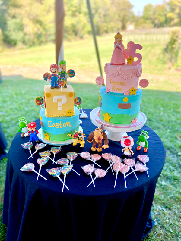 Party cake table with two, two-tiered cakes decorated with Super Mario Brothers decor. Suckers and figurines are scattered on the table around the cakes.