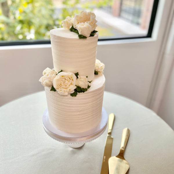 Two-tiered white wedding cake with floral decorations on a round table.